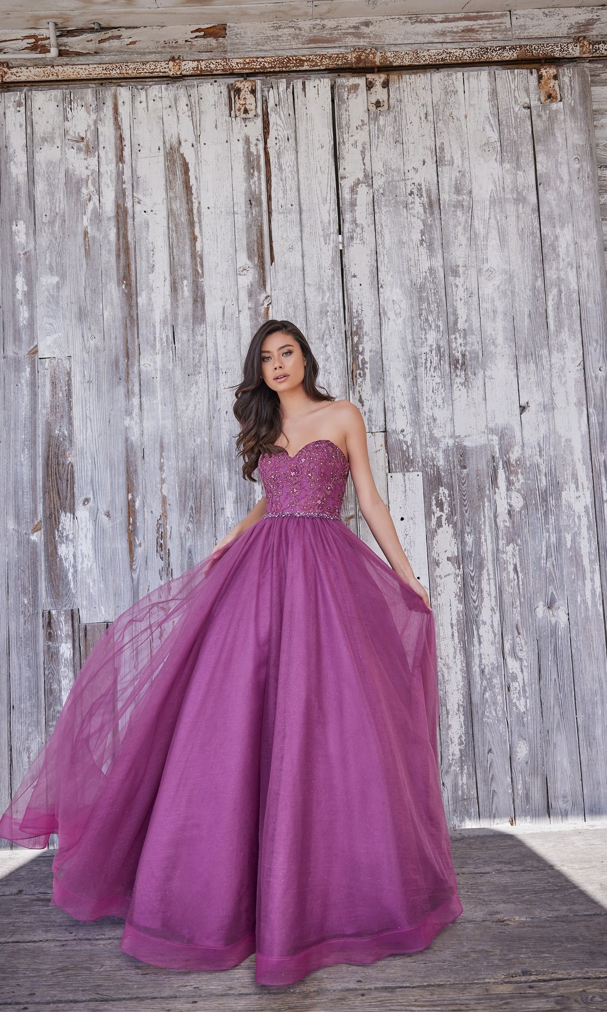 A woman in the Colette Strapless Sweetheart Prom Ball Gown CL5193 stands before a weathered wooden wall, gracefully holding out the sides of her purple beaded lace dress.